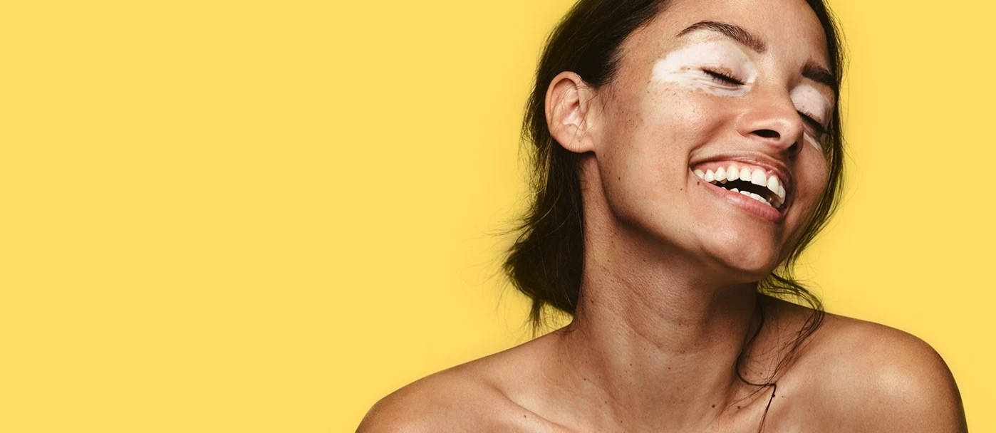 Mujer sonriente con crema sobre los pómulos, sobre un fondo amarillo.
